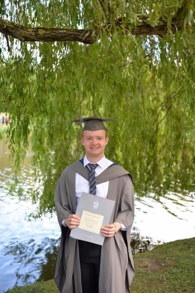Me in my graduation gown and cap, holding my degree under a willow tree